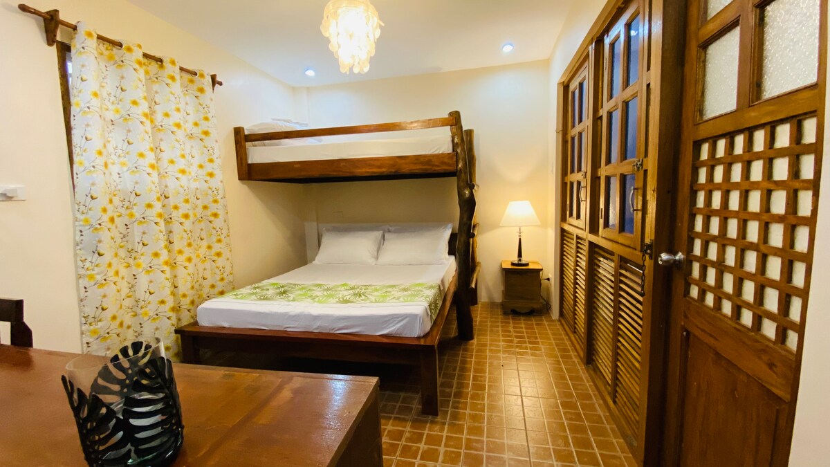 A compact sleeping area features two queen beds, one positioned above the other. A wooden dining table is visible, accompanied by a decorative centerpiece. Natural light enters through a window with wooden accents, while a bedside lamp provides additional illumination.