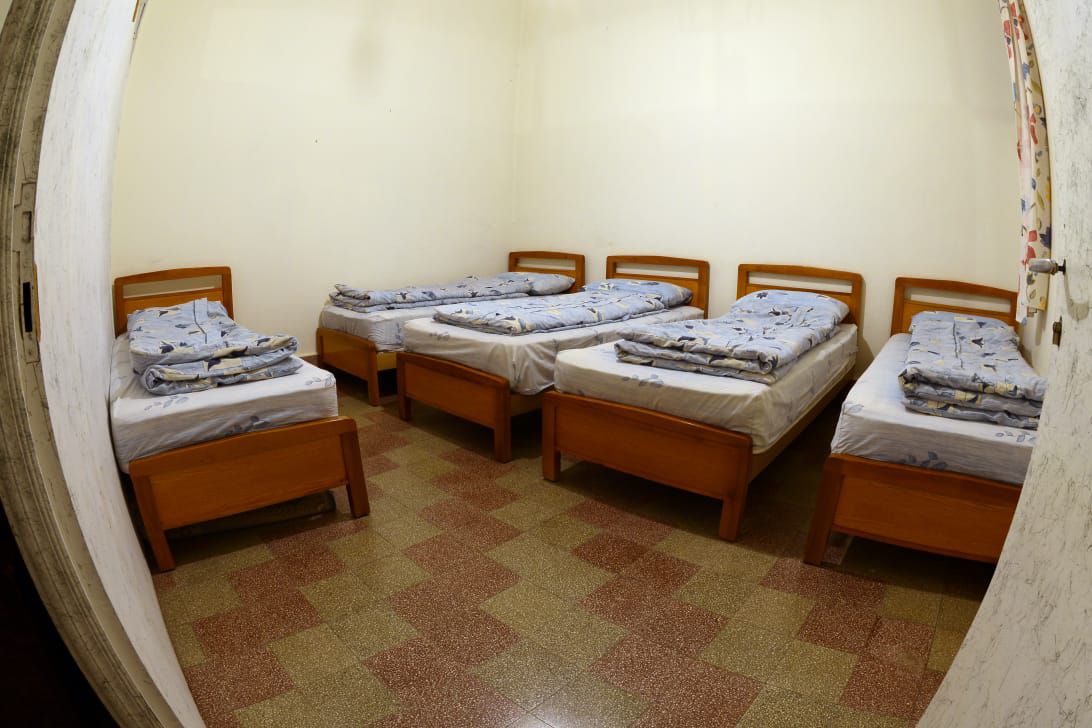 A spacious room contains four wooden beds arranged neatly. Each bed is dressed with decorative blue bedding. The floor features a patterned tile design in warm colors, and the walls are painted a light shade, creating a bright atmosphere.