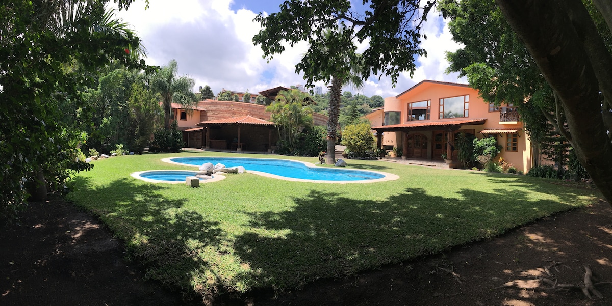 An expansive backyard features a curved swimming pool surrounded by well-maintained grass. Lush greenery and trees frame the space, offering shade. A large house with multiple windows is visible, complemented by a covered outdoor area, creating a peaceful setting ideal for relaxation.