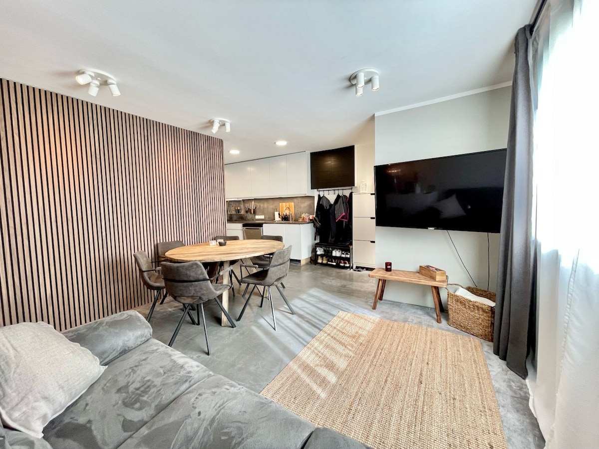 A modern living area features a cozy seating space and a dining table with six chairs, positioned next to a sleek kitchen. A large flat-screen TV is mounted on the wall, while natural light enters through the window, illuminating the room's neutral color palette.