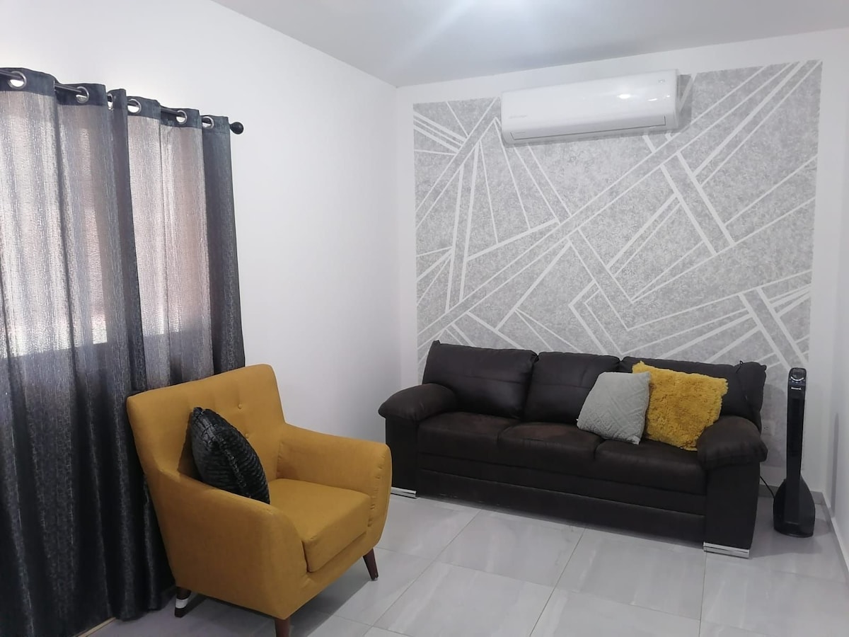 A modern living area features a dark sofa adorned with decorative pillows and a mustard yellow accent chair. Large black curtains frame the windows, while a patterned gray wall adds interest. An air conditioning unit is mounted above, ensuring comfort.