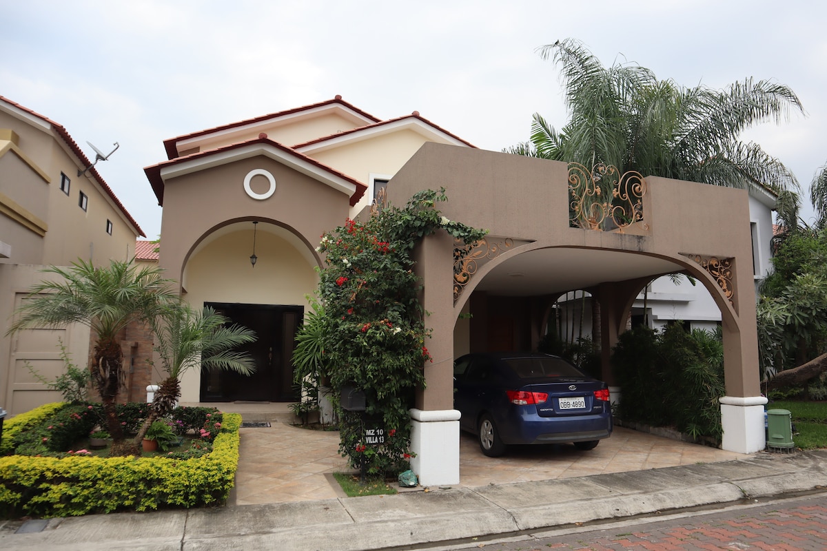The two-story house is framed by a well-maintained garden with colorful plants. A decorative carport provides shade for a parked vehicle, and a pathway leads to the inviting entrance. Lush palm trees enhance the tropical feel of the exterior.
