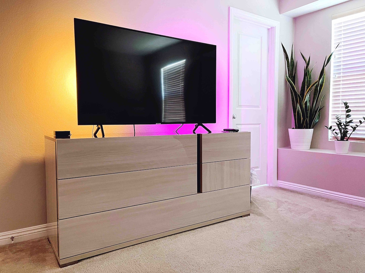 A modern TV is mounted on a sleek dresser with multiple drawers, positioned against a soft-hued wall. Natural light filters through a nearby window adorned with blinds, while green plants are placed in pots, adding a touch of nature to the space.