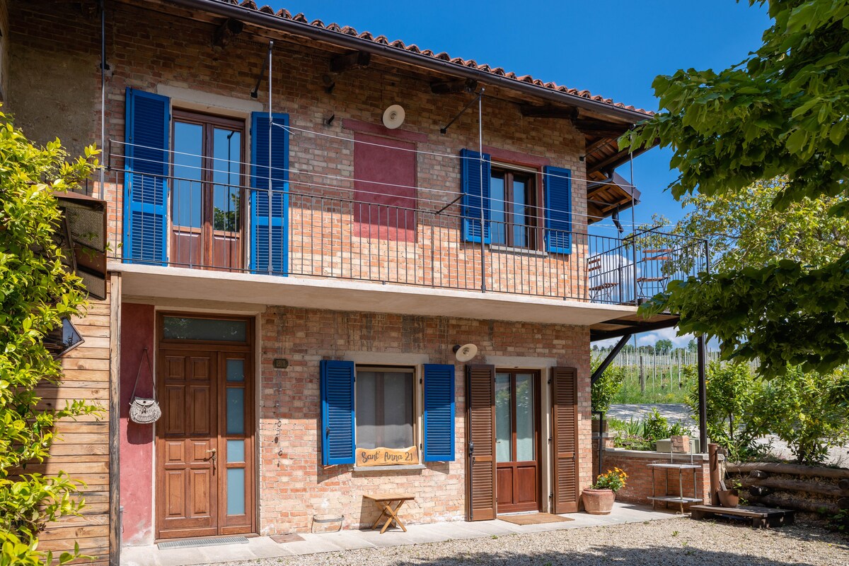 A two-story, rustic brick house is shown, featuring blue shutters and a welcoming entryway. A small balcony is visible on the upper level, complemented by a cozy outdoor seating area. The surrounding greenery and vineyard view enhances the natural setting.
