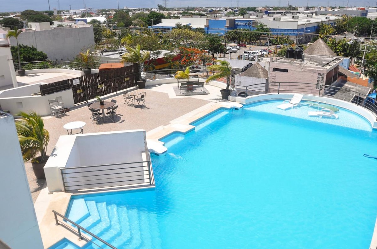 A rooftop pool area features a spacious swimming pool with steps leading down into the water. Lounge chairs and small tables are arranged nearby, surrounded by tropical plants. A scenic view of the city is visible in the background.