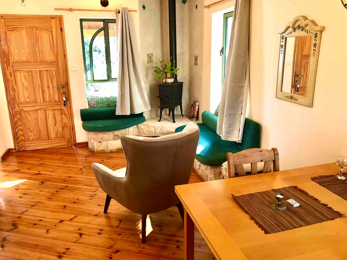 A cozy seating area features two green benches and a comfortable armchair, arranged around a small wood stove. Natural light streams through the windows, illuminating the warm wooden floor and highlighting the inviting atmosphere of the space.