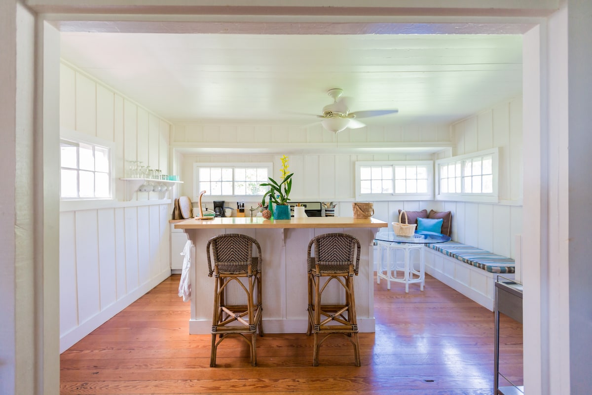 A bright kitchen area is visible, featuring a large island with seating for two. Light wood accents and open windows create a spacious feel. A small nook with cushions adds a cozy touch, while several glassware items are neatly arranged on the shelves.