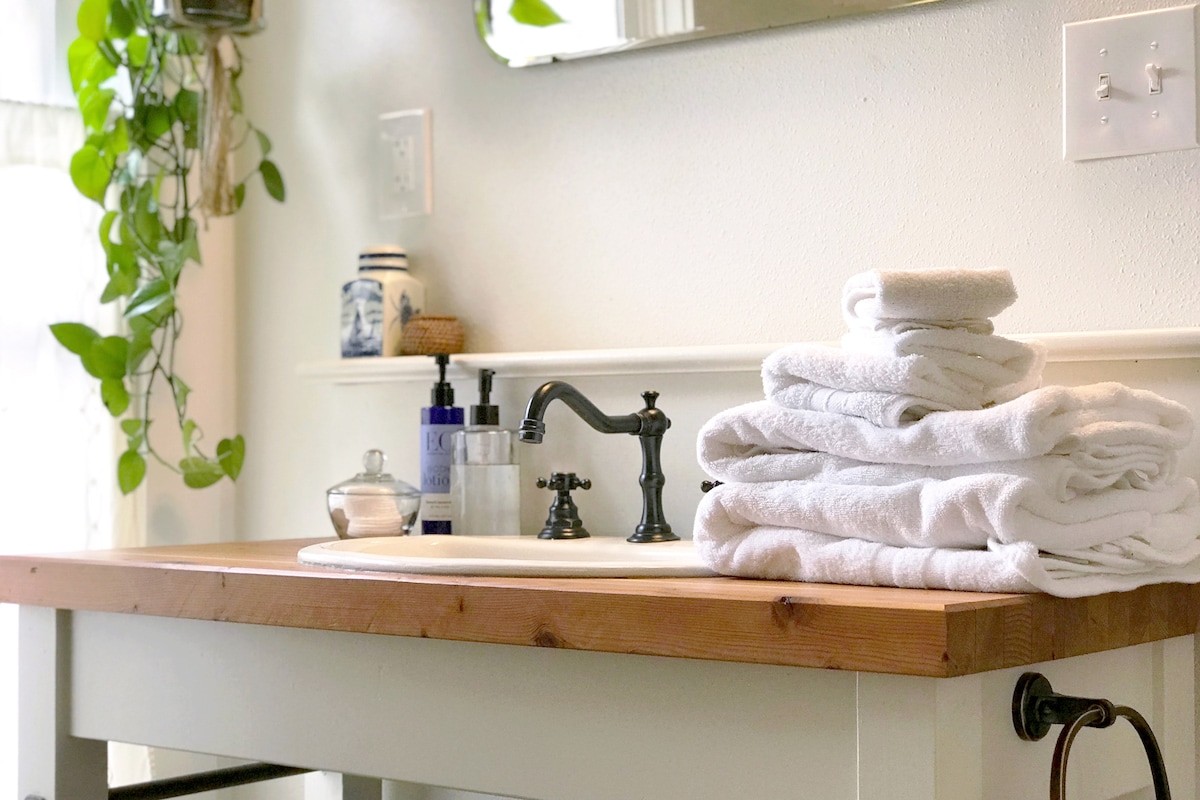 A bright bathroom features a rustic wooden countertop and a white sink. Freshly laundered white towels are neatly stacked beside the sink. Decorative elements include a small plant and an elegant mirror reflecting natural light.