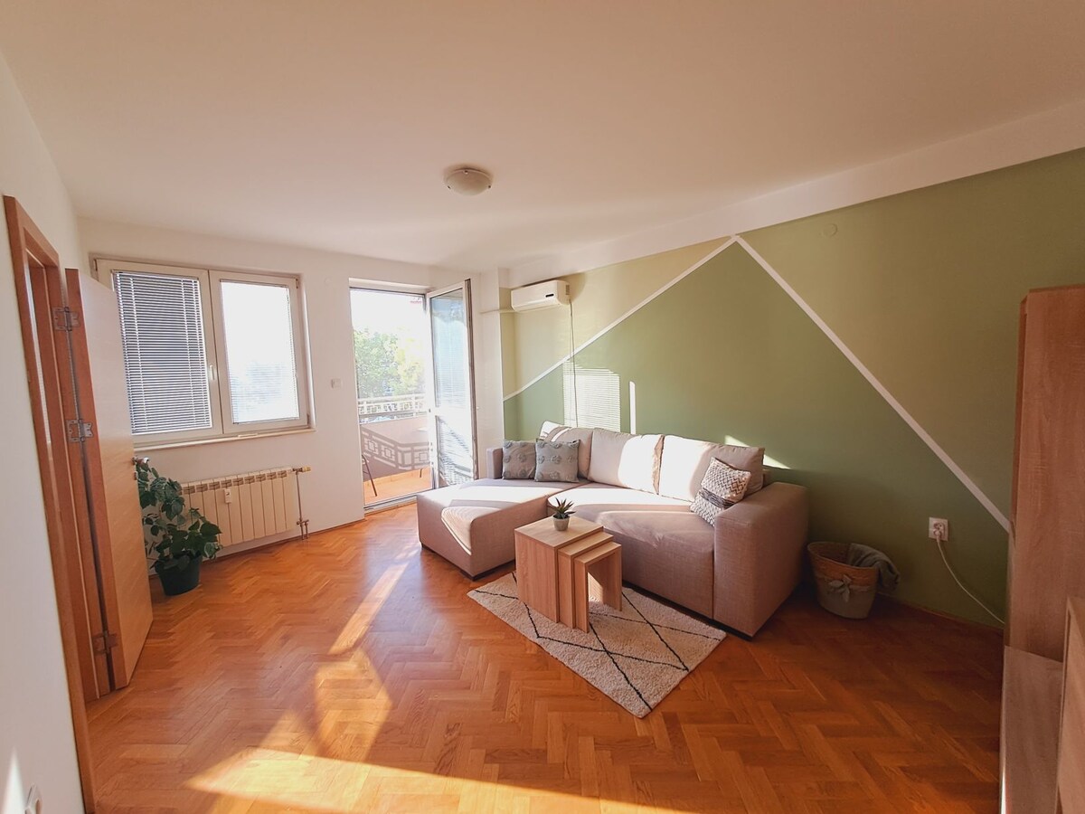 A spacious living area features a light-colored sofa paired with a wooden coffee table. Natural light floods through large windows, illuminating the room's modern design. A small plant adds a touch of greenery, creating a relaxed environment. The walls are accented with a soft green hue.