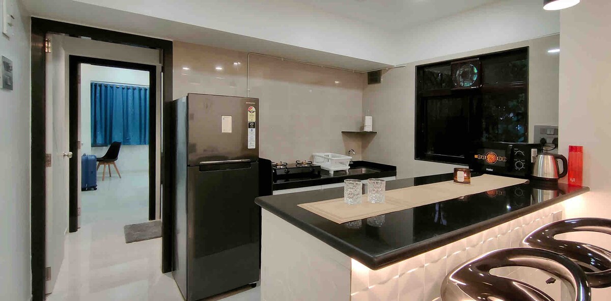 The kitchen features a sleek design with modern appliances, including a refrigerator and microwave. A gas stove is positioned against the wall, accompanied by basic utensils and cutlery arranged on the counter. Large windows provide natural light, enhancing the cooking area.