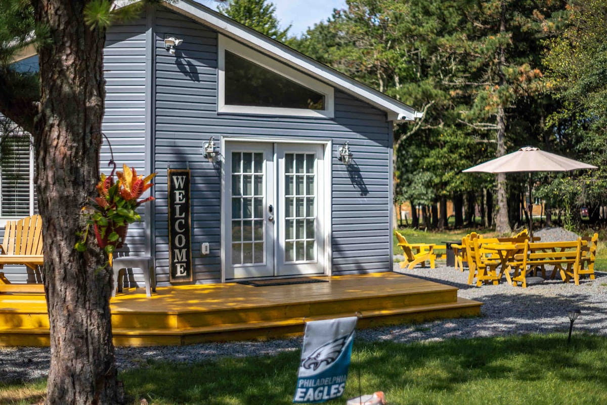 A charming exterior view of the lodge is displayed, featuring a welcoming sign above double French doors. A spacious deck leads to outdoor seating arrangements, including a large umbrella. Surrounding greenery complements the inviting atmosphere of the outdoor space.