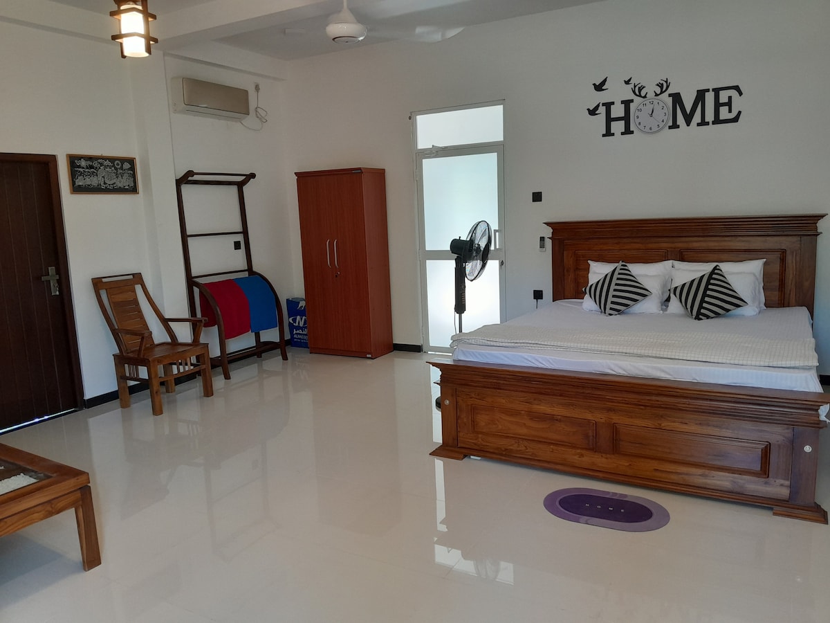 The private room features a spacious wooden bed dressed in crisp white linens, complemented by decorative pillows. A wardrobe and a fan are visible, while a comfortable wooden chair is positioned next to an accent table. Natural light enters through the window, enhancing the room's bright interior.