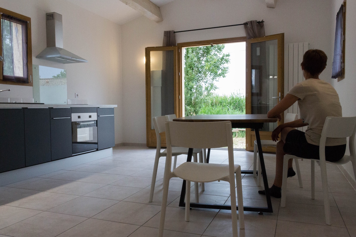 The kitchen area features modern cabinetry and stainless-steel appliances, with a dining table positioned near large glass doors. Natural light floods the space, illuminating the tiled flooring. A young person is seated at the table, gazing out towards greenery visible through the open doors.