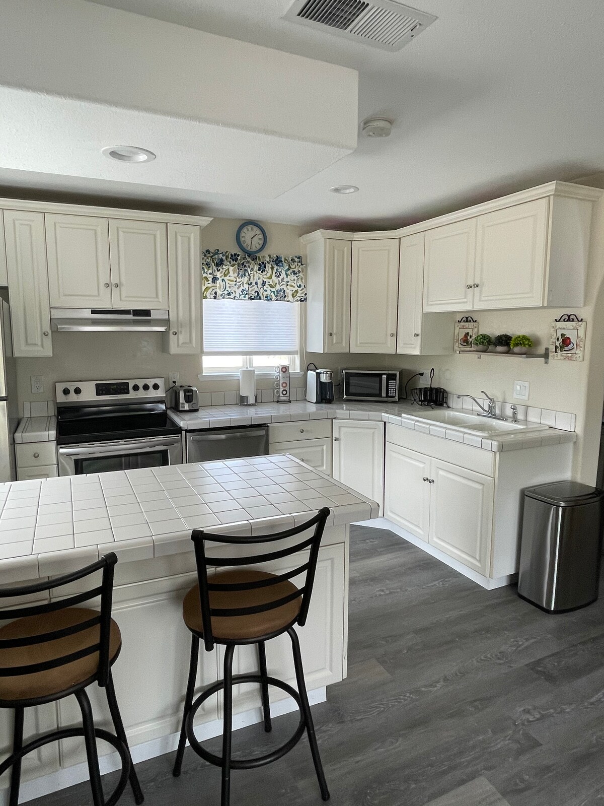 The kitchen features white cabinetry and a tiled countertop. Stainless steel appliances, including an oven and microwave, are present. Two barstools are positioned at the counter, and natural light enters through the window adorned with a floral valance.