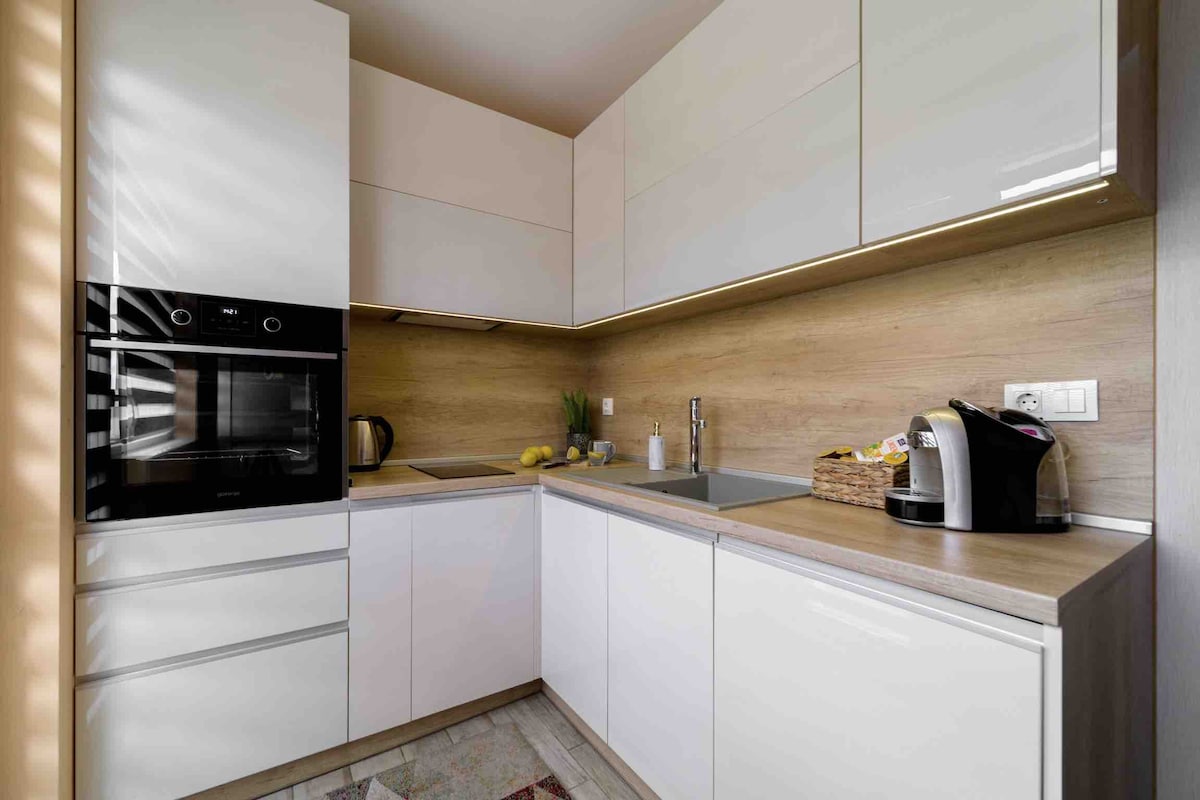A modern kitchen featuring sleek white cabinetry and a wooden countertop. Built-in appliances, including an oven and a kettle, complement the minimalist design. A coffee machine and a basket of fresh fruits add practical touches, while natural light filters through the space.