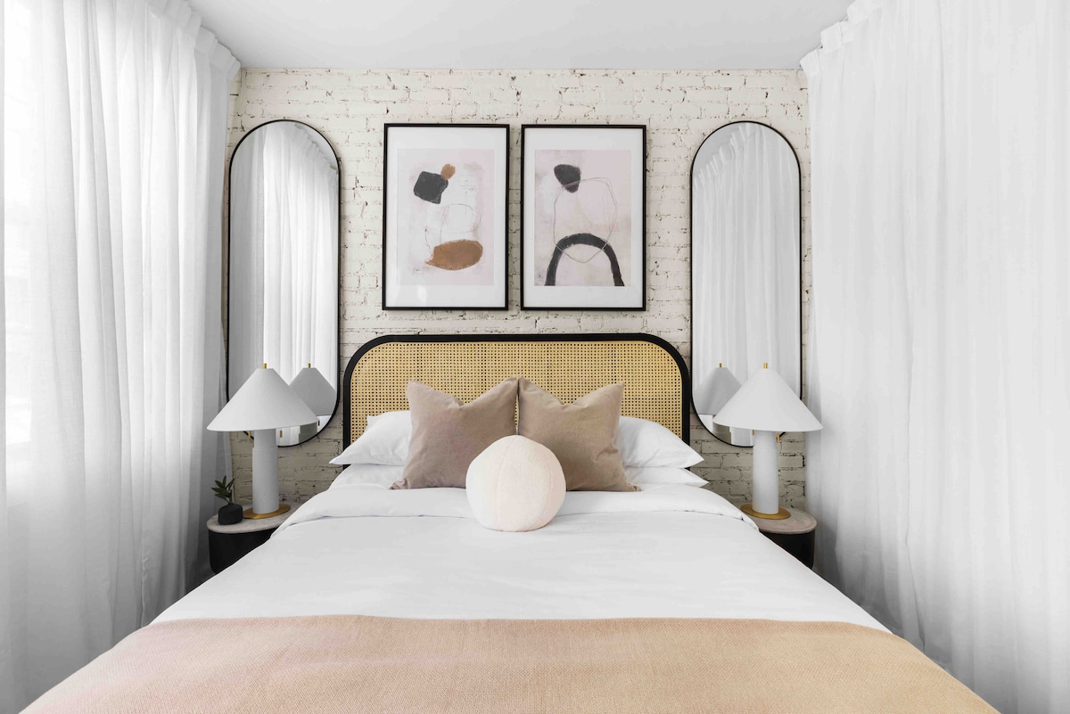 The bedroom features a stylish bed with a light-colored blanket and plush pillows. Two round mirrors and framed artwork adorn the wall, complemented by elegant table lamps on either side. Sheer white curtains frame the windows, adding a soft touch to the modern decor.