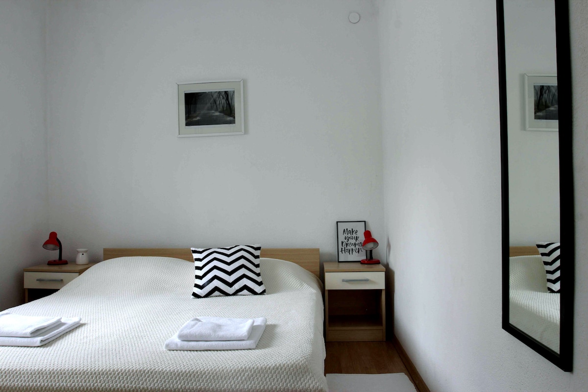A bright bedroom features a spacious bed with a light-colored coverlet and an accent pillow. Two bedside tables with lamps are positioned on either side. A large mirror reflects the room, and a framed black-and-white photo adorns the wall, contributing to a simple, modern aesthetic.