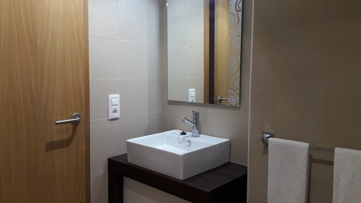 A modern bathroom features a white square sink positioned on a dark wooden vanity. Above, a large mirror reflects the space, while a hand towel hangs neatly on a metal rack to the right. Neutral tiles create a clean and simple backdrop.
