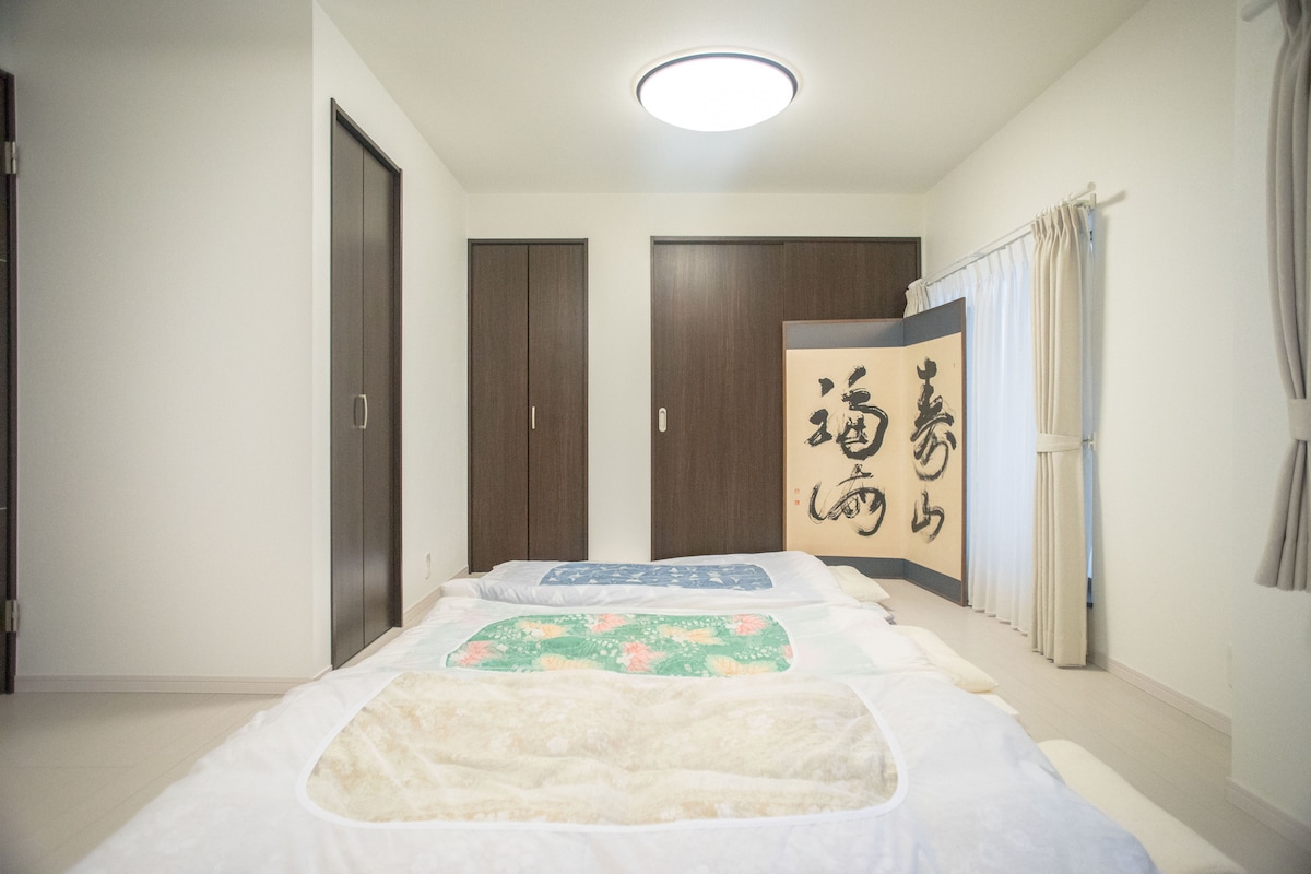 A spacious room features multiple futons laid out on a light-colored floor. Sliding wooden doors provide access to storage, while a decorative screen with calligraphy adds cultural charm. Natural light filters in through a window, enhancing the airy feel of the space.