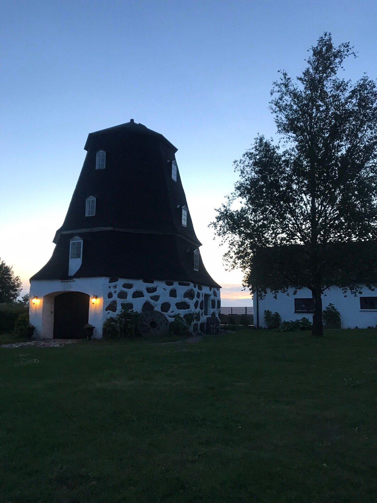 A charming, traditional building with a distinctive dark roof structure is showcased in the evening light. The stone facade is paired with a white structure nearby. Lush greenery surrounds the area, with a clear sky transitioning to twilight.