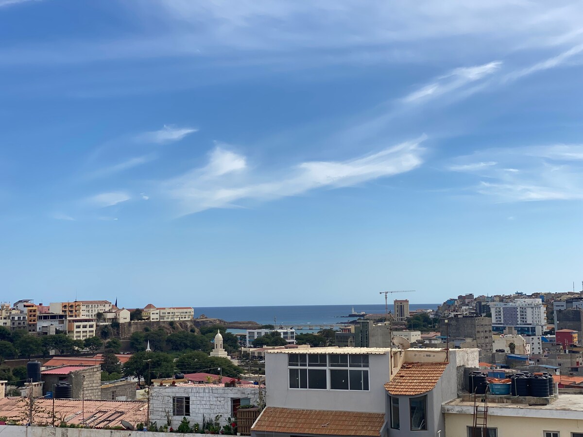 A panoramic view captures coastal scenery with the ocean stretching towards the horizon. Various buildings, including low-rise structures with terracotta roofs, populate the foreground. A clear sky with soft clouds enhances the tranquil atmosphere of the surrounding area.