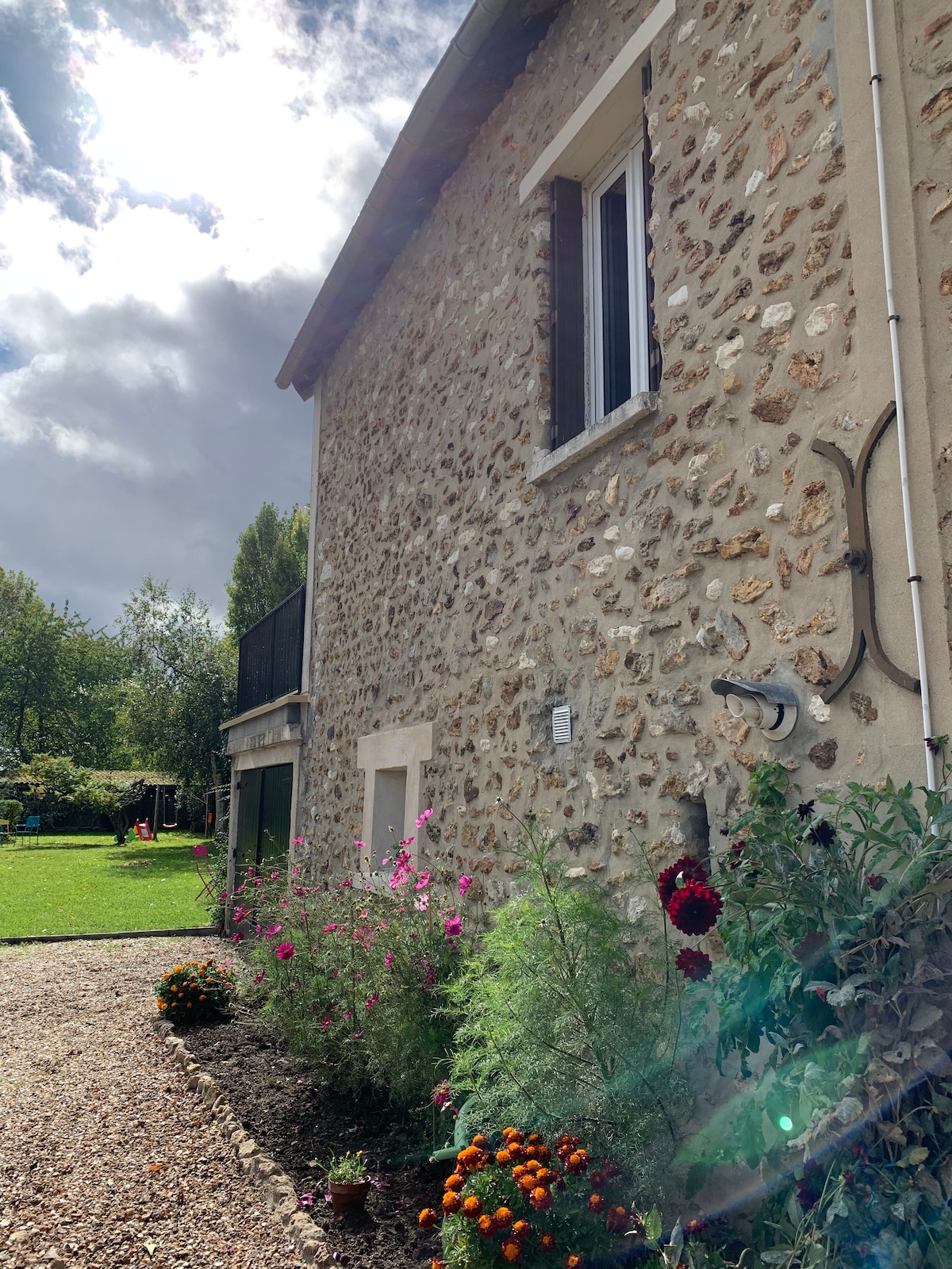 The exterior of the house is showcased, featuring a rustic stone facade complemented by blooming flowers in vibrant colors. A spacious green yard is visible in the background, enhancing the natural surroundings. Large windows are framed by greenery, inviting light into the home.