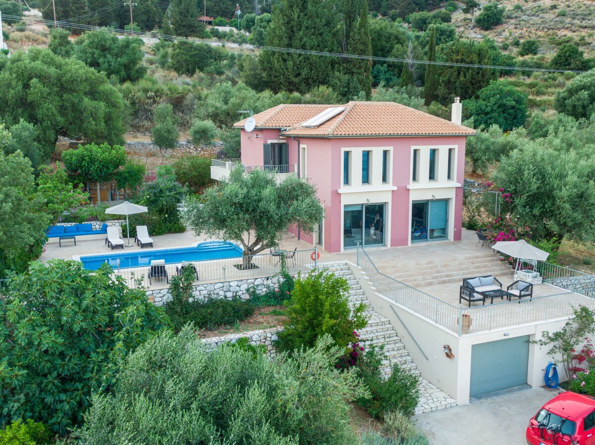 A two-story villa is shown, surrounded by lush greenery and olive trees. A spacious pool area is visible, featuring sun loungers and umbrellas. The exterior is painted in soft pink tones, and a patio is set with a table and chairs. A garage is accessible at the ground level.