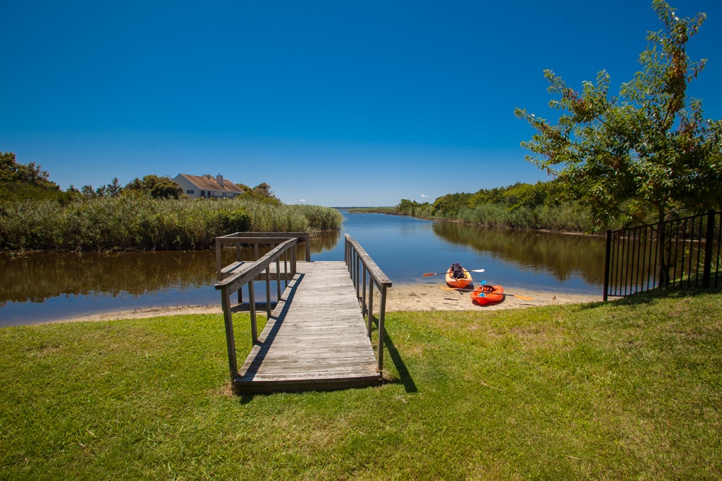 A wooden dock extends over a tranquil inlet, bordered by lush greenery. Two kayaks in vibrant colors rest on the sandy bank, inviting exploration. The clear blue sky overhead enhances the serene surroundings, creating a peaceful outdoor retreat.
