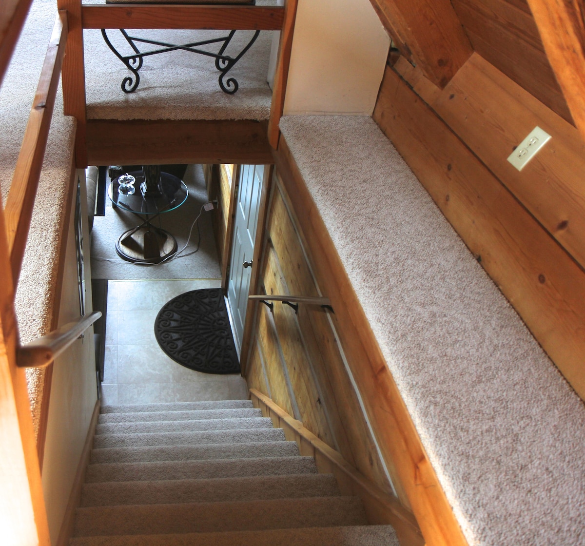 A view from the loft shows a staircase leading down to the main living area. The carpeted stairs are lined with wooden walls and a handrail, while a round glass table is visible at the bottom of the stairs. Soft lighting gently illuminates the space.