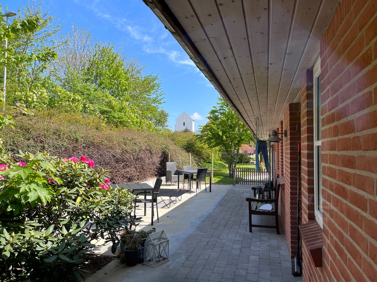 A brick patio extends alongside lush greenery and vibrant flowers. Outdoor seating arrangements, including a table and chairs, are visible in a shaded area. A pathway leads towards an open garden space under a clear blue sky, inviting moments of relaxation and connection with nature.