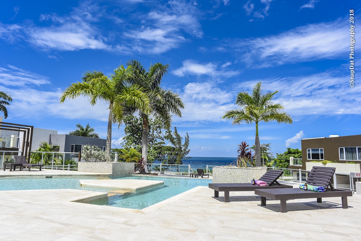 A spacious pool area is surrounded by palm trees, featuring lounge chairs with towels draped over them. The sky is bright with scattered clouds, and the ocean is visible in the background, creating a serene outdoor environment.