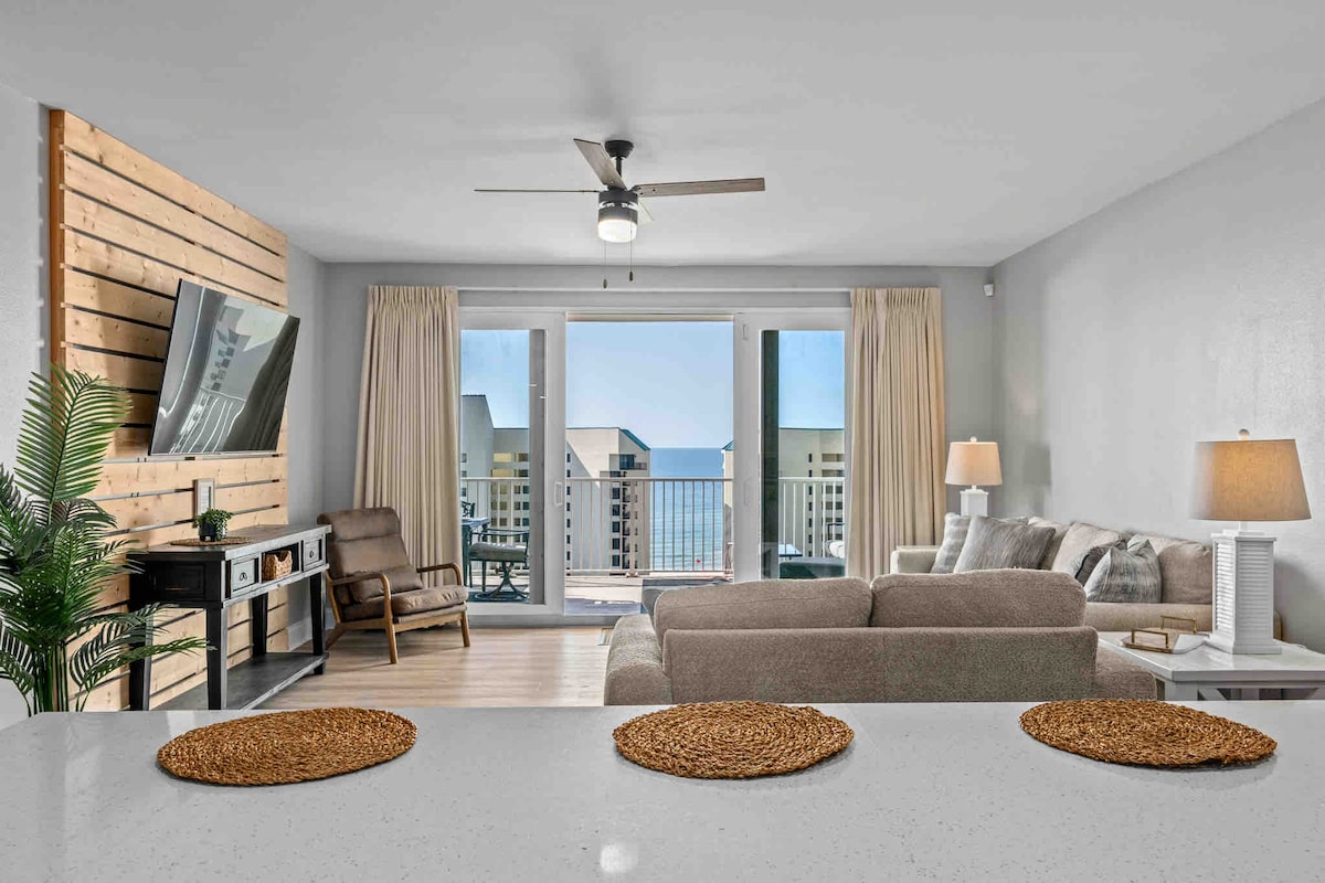 A bright living area is framed by large sliding glass doors that offer expansive views of the ocean. A cozy seating arrangement includes a light-colored sofa and an armchair. A neutral-toned wall enhances the aesthetic, while a wooden accent wall adds texture to the space.