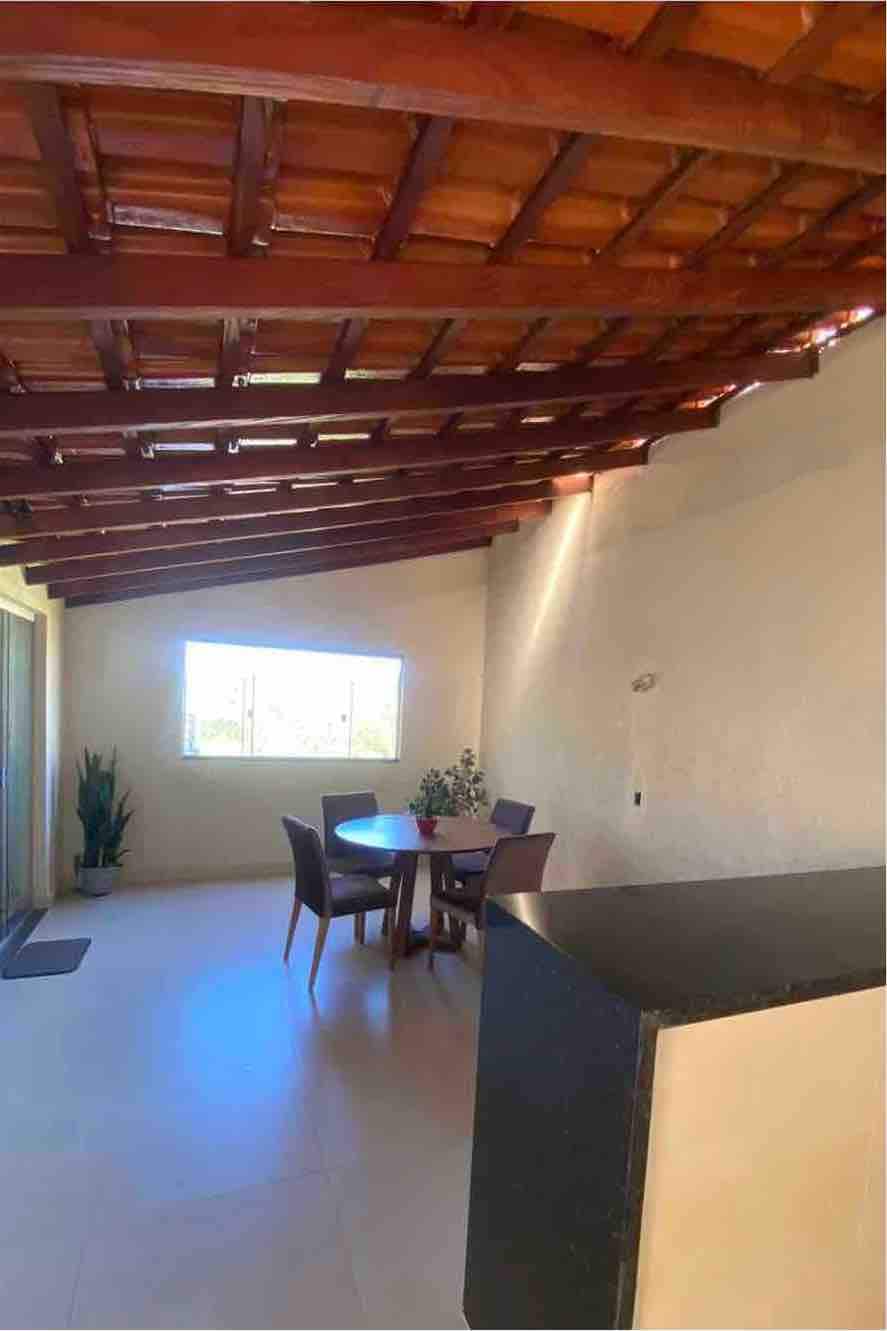 A sunlit area is shown, featuring a wooden ceiling with exposed beams. A dining table with four chairs is positioned near a window, allowing natural light to fill the space. Green plants add a touch of nature, and a dark countertop is partially visible in the foreground.