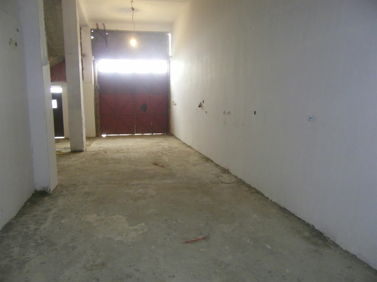 A spacious garage area is depicted, featuring an unfinished concrete floor and plain white walls. Light enters through a large red door at the far end, providing a sense of openness. Minimal elements are visible, offering a blank canvas for storage or vehicle parking.