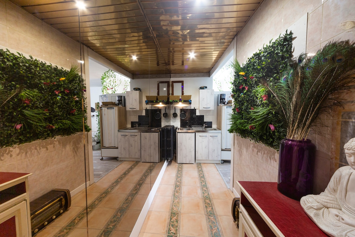 The kitchen features a stylish design, with greenery accented on the walls. Contemporary cabinetry and appliances are present, complemented by natural light from nearby windows. A hallway leads to the kitchen area, showcasing tiled flooring that extends through the space.