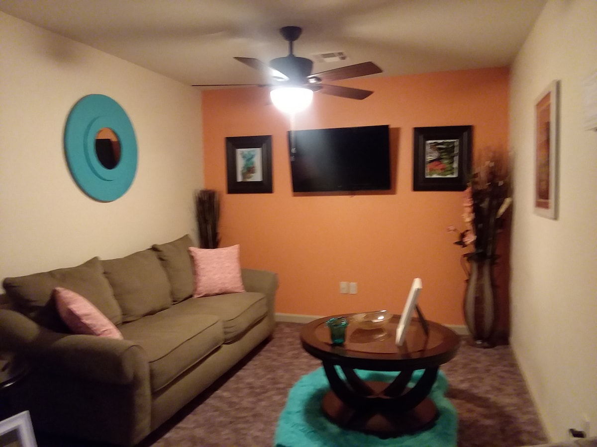 A cozy living area features an orange accent wall contrasted by neutral furnishings. A sectional sofa with pink accent pillows is arranged around a circular coffee table. A ceiling fan provides airflow, and wall art adds a touch of color to the space.