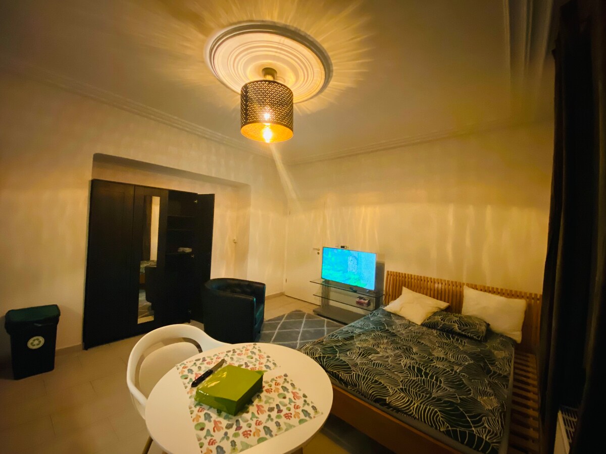 A modern room features a spacious bed adorned with a floral-patterned blanket. A circular dining table sits nearby, accompanied by two chairs. A television is positioned on a glass stand. Soft lighting creates a warm ambiance, while dark curtains frame a window.