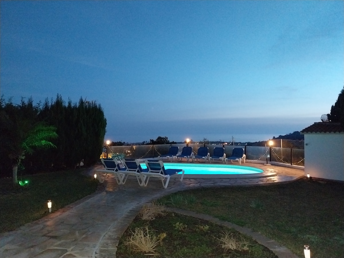 A serene evening setting is depicted with a softly illuminated pool surrounded by lounge chairs. The garden is bordered by lush greenery, with pathway lights creating a welcoming ambiance. A distant view hints at the horizon, enhancing the tranquil atmosphere.