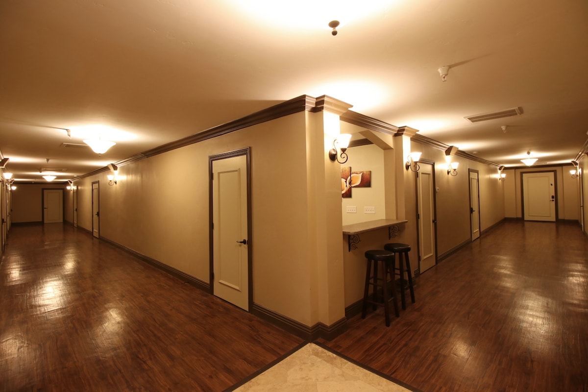 A well-lit hallway is shown, featuring smooth wooden floors and warm-toned walls. Several light fixtures provide illumination, while doors line both sides. A small seating area with a stool is positioned against one wall, creating a functional and inviting space.