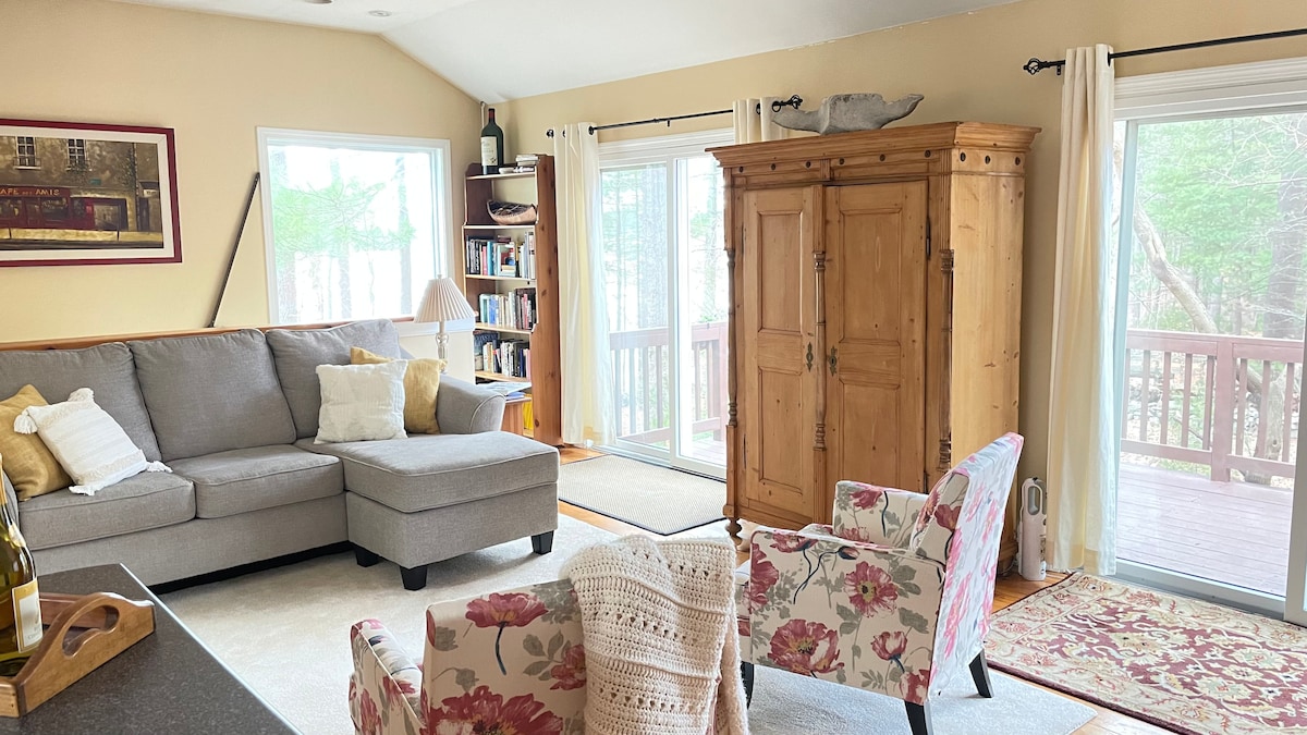 A comfortable living area features a sectional sofa in light gray, complemented by floral armchairs. A wooden armoire stands against the wall, while natural light floods in through large sliding doors leading to a deck outside. Bookshelves are visible in the background.