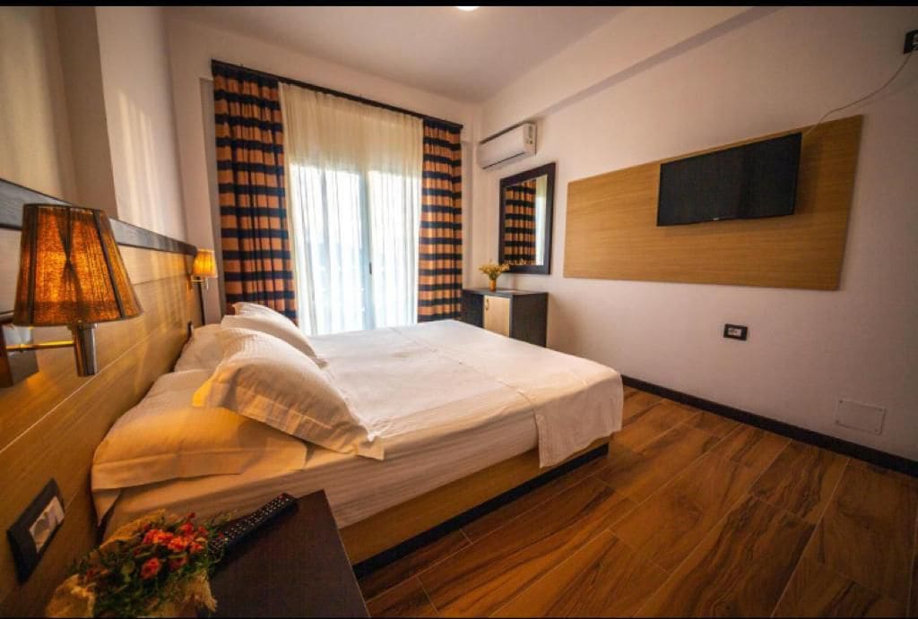 A well-appointed sleeping area features a spacious bed with crisp white linens and a soft headboard. A large window allows natural light to fill the room, framed by striped curtains. A flat-screen TV is mounted on the wall, and a small decorative arrangement sits on the bedside table.