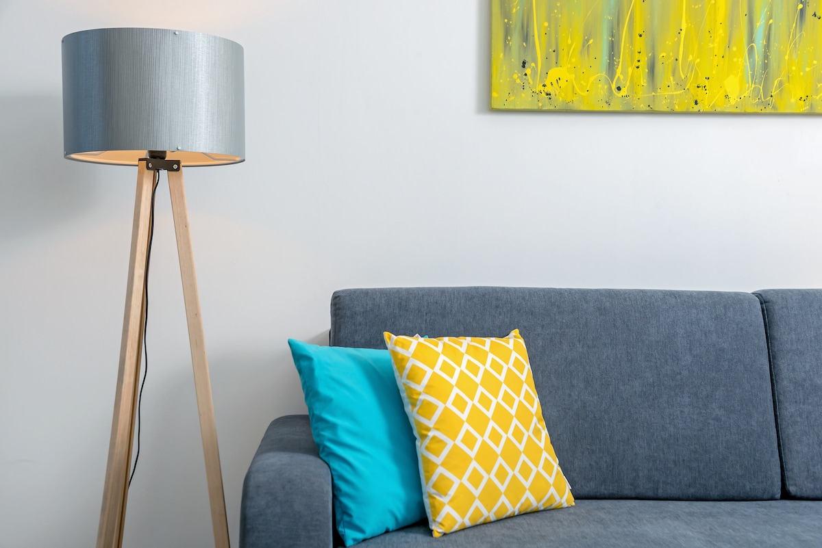 A modern living area is featured with a gray sofa adorned with two decorative cushions, one in a vibrant yellow pattern and the other in a solid turquoise. A tall, wooden lamp with a silver shade provides soft illumination, while a bright abstract painting in yellow hangs on the wall.