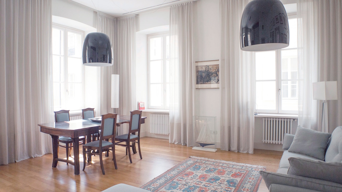 An airy living area features a dining table surrounded by six chairs and a comfortable sofa. Tall windows are complemented by light, flowing curtains, allowing natural light to fill the space. A colorful rug adds warmth beneath the furniture.