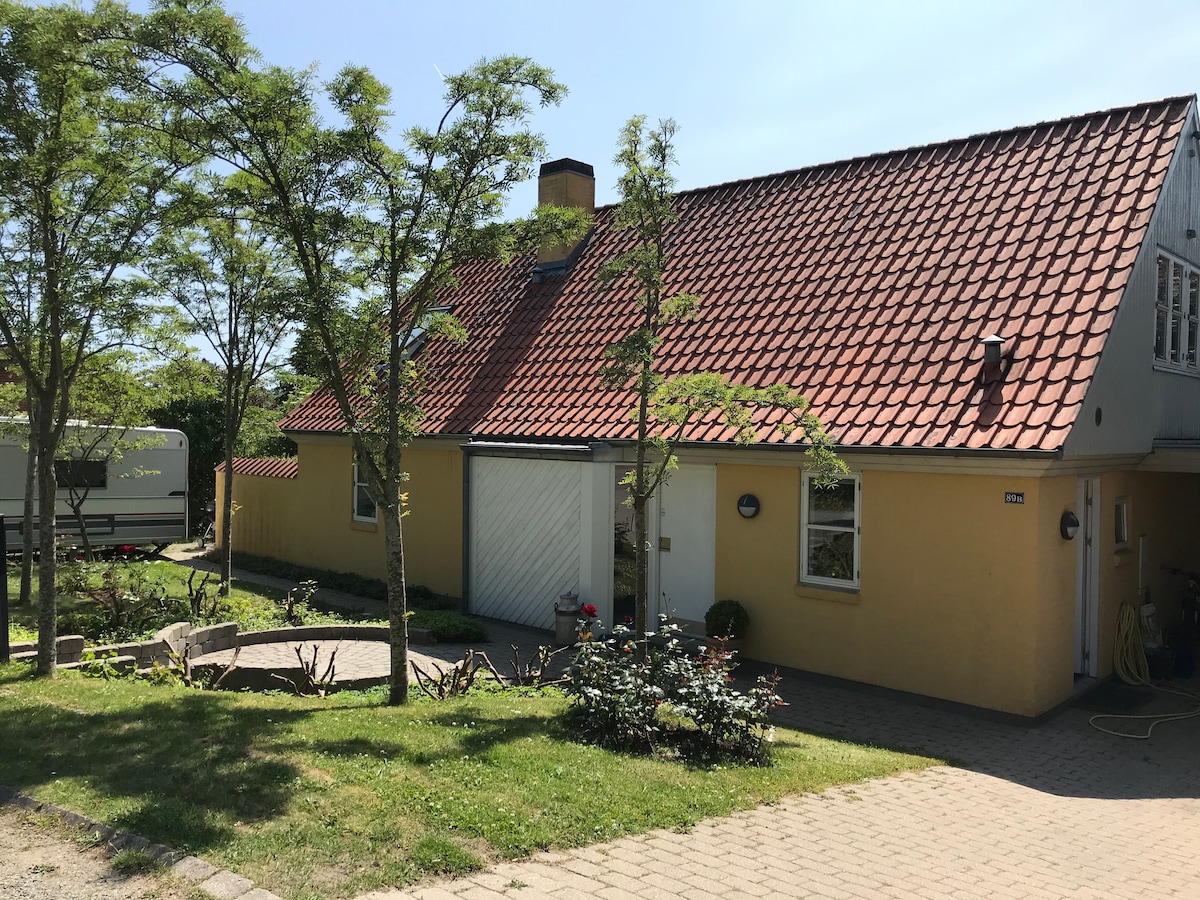 A charming house is set among trees and greenery, featuring a sloped roof with red tiles. A paved path leads to the entrance, while a small seating area with natural landscaping is visible in the front yard. The sunny scene conveys a peaceful ambiance.