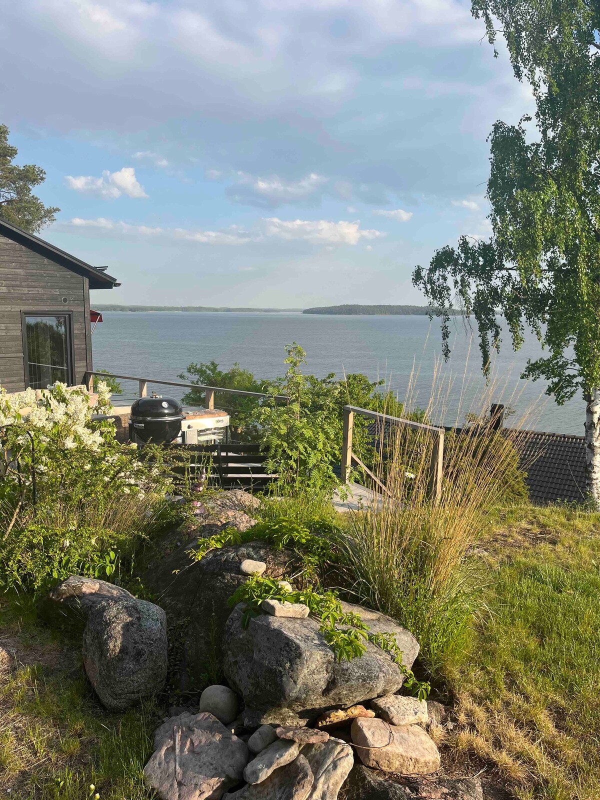 A scenic view showcases the exterior of the guest cabin set against a backdrop of Bråviken. Lush greenery and a rock formation lead to a wooden deck with a barbecue grill, overlooking the serene water. Gentle waves reflect the sky's soft hues.