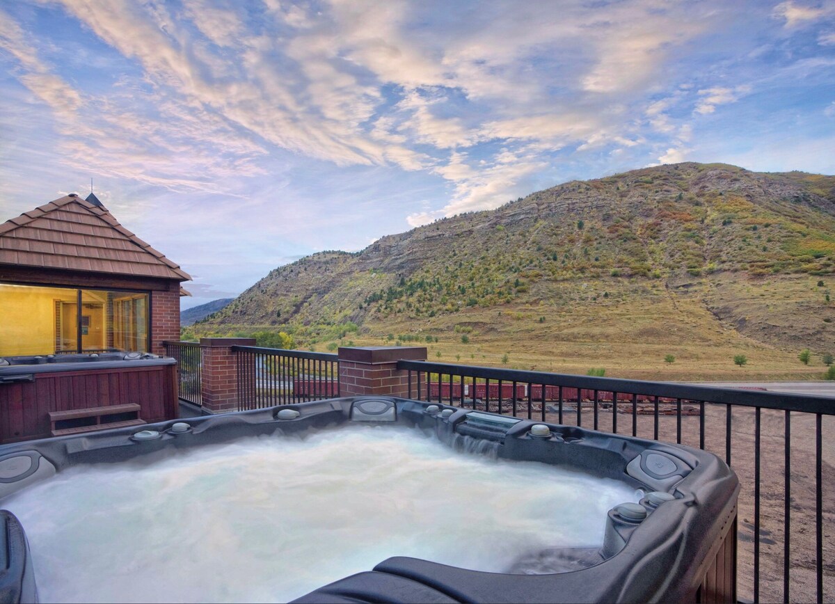 An outdoor hot tub is positioned on a deck, overlooking rolling hills and a scenic landscape. The surrounding railing offers safety while maintaining open views of the natural beauty. The sky showcases a mix of soft clouds and hues during sunset.