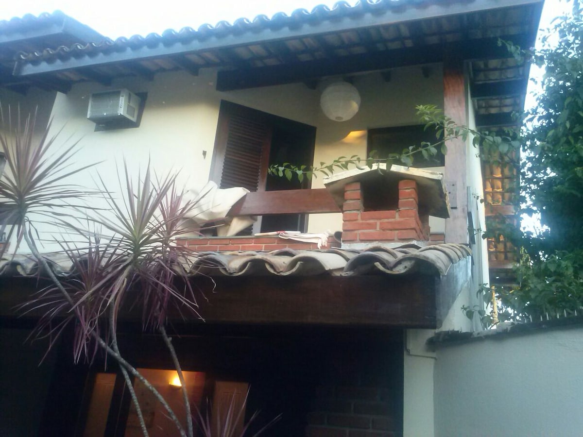 The exterior of a two-story house is visible, featuring a tiled roof and wooden beams. Lush green plants frame the entrance, and an air conditioning unit is mounted on the wall. Soft lighting is seen through the windows, hinting at the cozy interior.