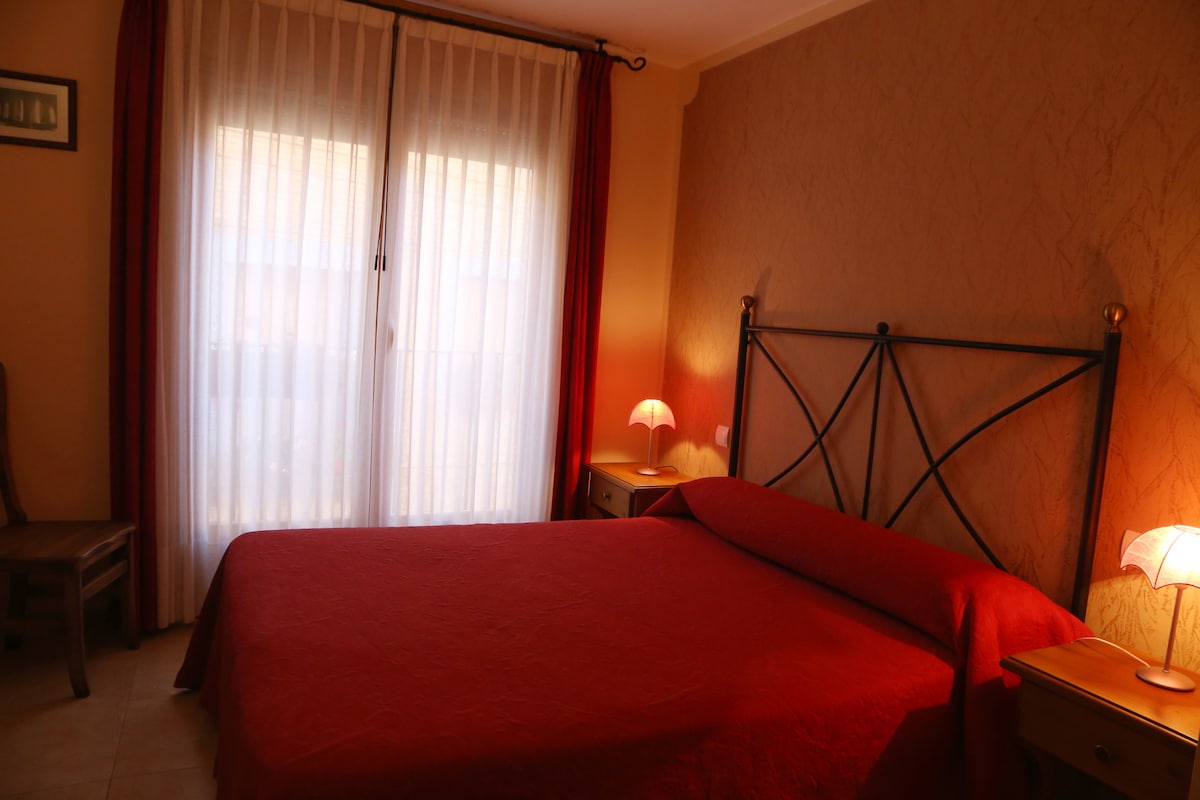 A warm bedroom features a large bed with a red coverlet. Soft lighting from two bedside lamps creates a cozy ambiance. Two large windows draped with sheer curtains allow natural light to fill the room while maintaining privacy.