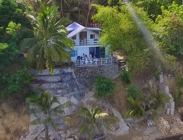 A charming beach house is nestled on a cliffside, surrounded by lush palm trees. A spacious balcony overlooks the crystal-clear waters below, providing a serene escape. The house is partially shaded by trees, enhancing the natural coastal setting.