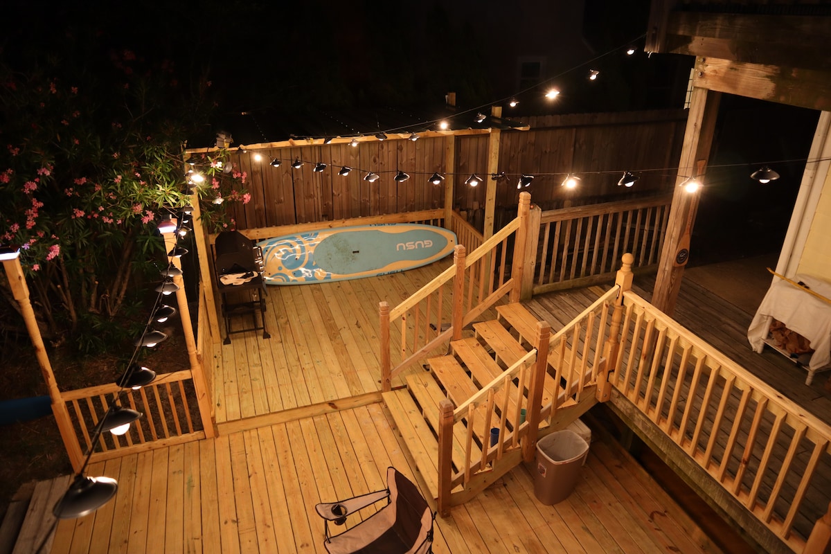 A wooden deck area is illuminated by string lights, providing a warm glow in the evening. A large paddleboard rests against the railing, with a chair nearby. The space is bordered by lush greenery and a wooden fence, creating a cozy outdoor setting.